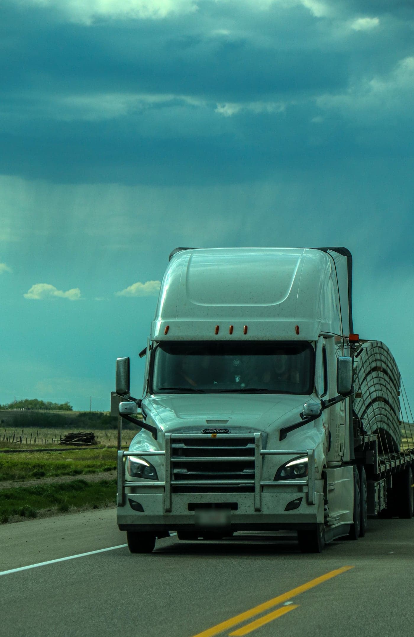 White Freightliner semi-truck approaching on a two-lane highway under a stormy sky